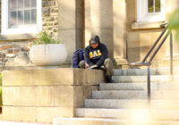 A university student in a DAL hoodie sits on the steps of a stone building on a university campus, writing in a notebook.
