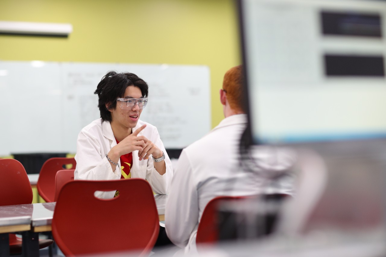 Two people are seen talking, both wearing lab coats, sitting at a table with red chairs.
