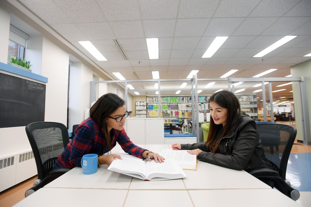 A person with long black hair is sitting holding open a book as another person across a table is sitting.