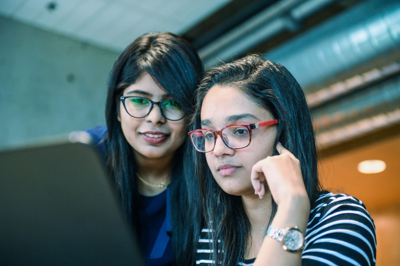 Two people wearing glasses with long black hair are looking at a computer screen.