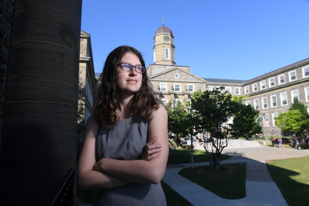 A person wth long brown hair and glasses wearing a grey sleeveless dress has their arms folded in front of the Henry Hicks building.