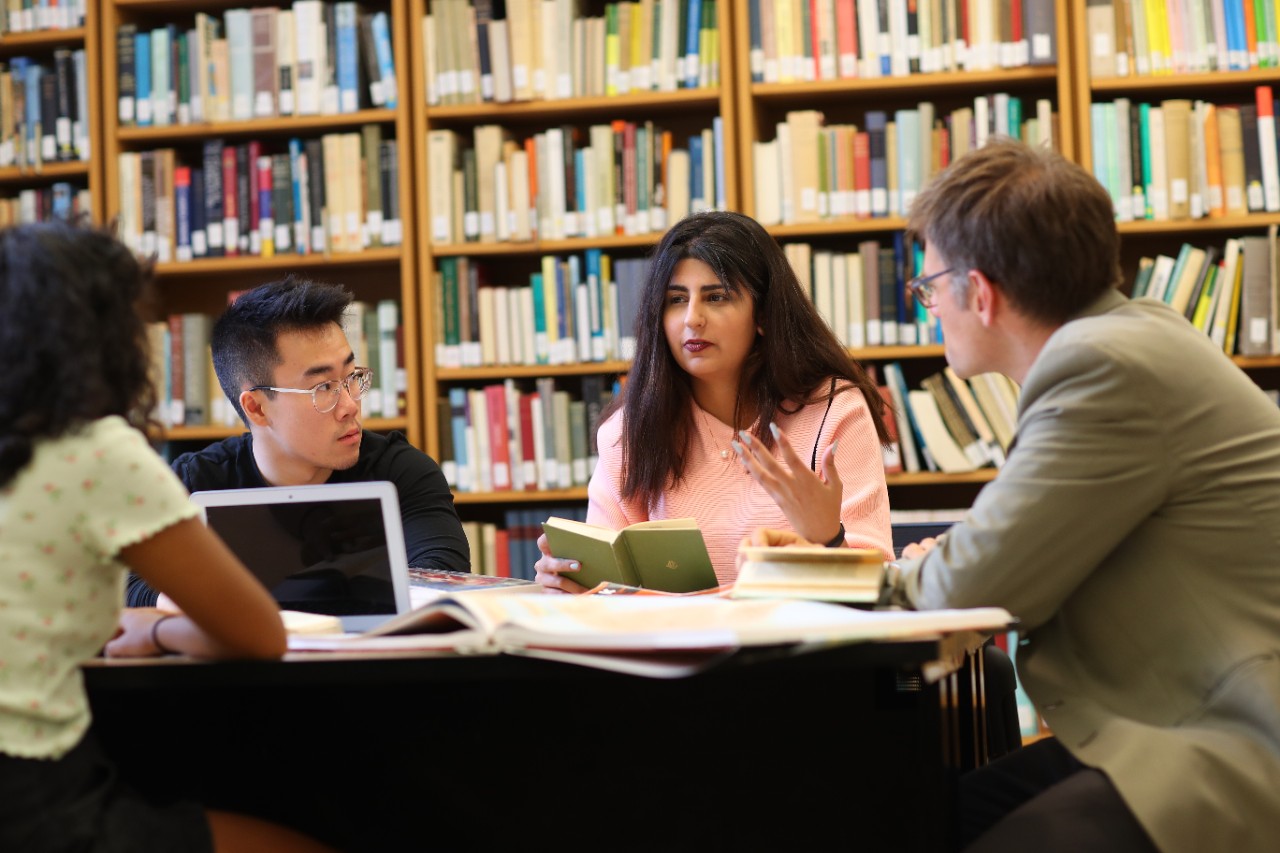 Four students sit around a desk with books and a laptop with stacks of books behind them.
