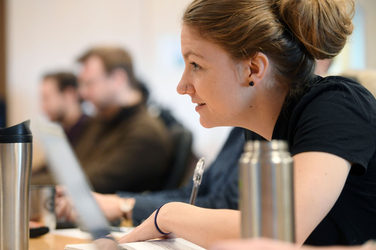 A person in profile wearing a shortsleeve black shirt is sitting at a desk using a notebook paying attention to someone off-camera.