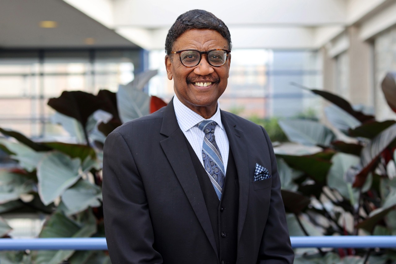 A portrait of Ruck smiling and dressed formally indoors with greenery in the background.