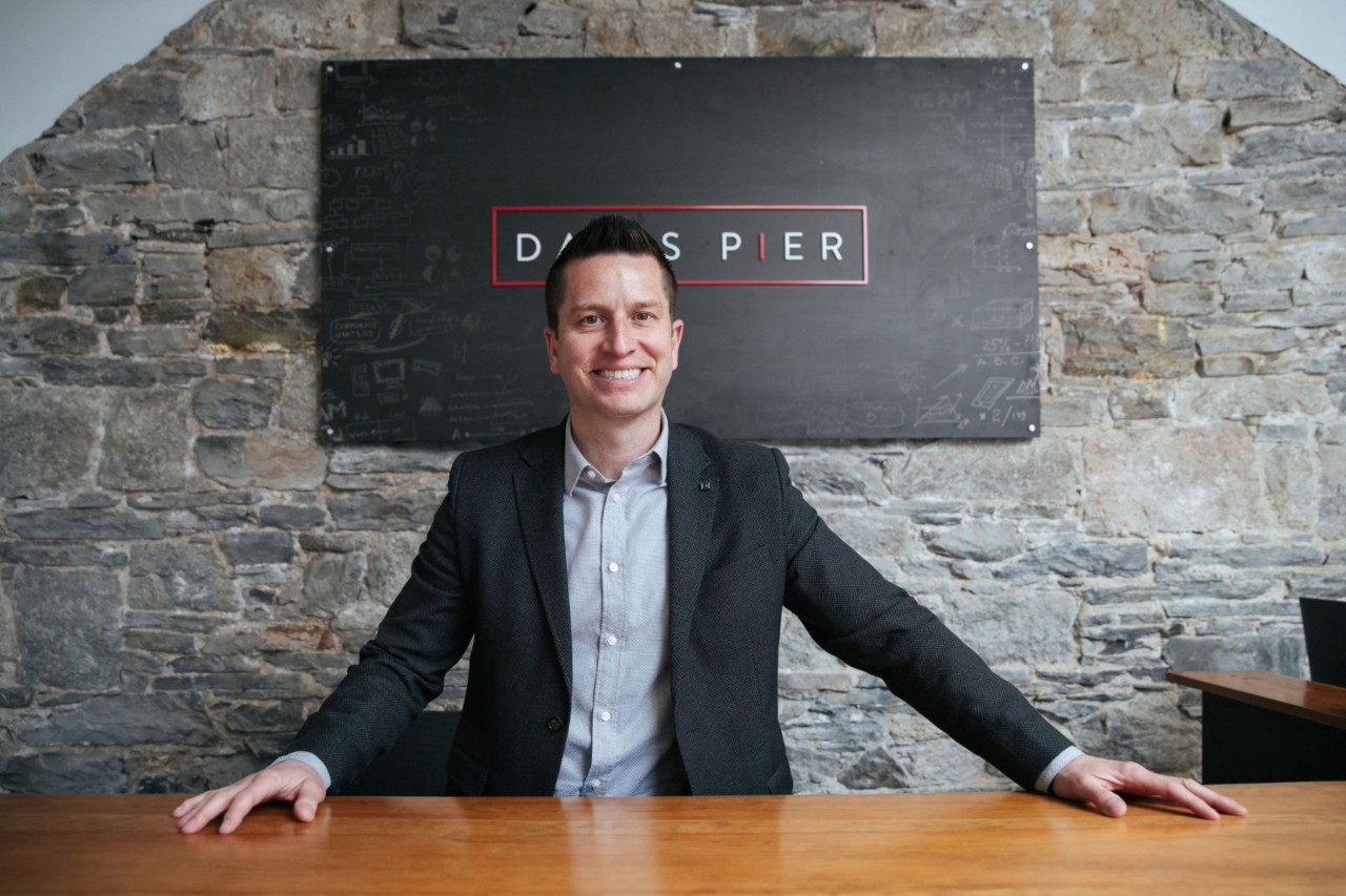 Mike Davis is smiling with his arms outstretched on the wooden desktop that he stands behind with a stone wall behind him..