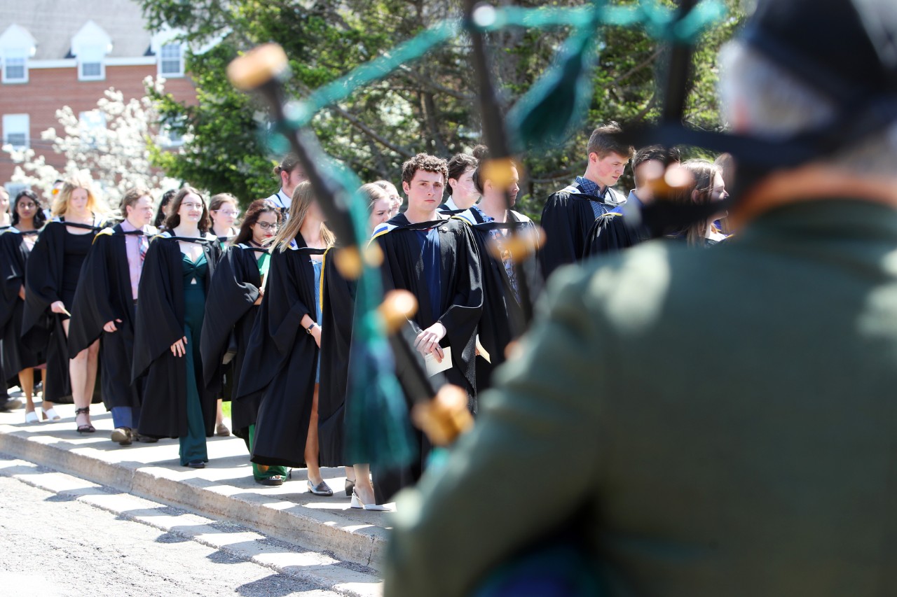 A line of students wearing black graduation gowns is seen through the pipes of a bagpiper.