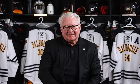 A man wearing seated in a hockey locker room with hockey jerseys on display in the background