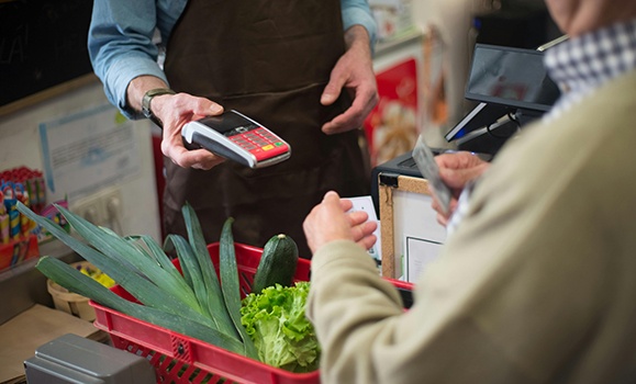 A close up shot of a cashier holding a payment terminal out to a customer who is purchasing groceries. In his basket are fresh vegetables - lettuce, cucumber and green onion.