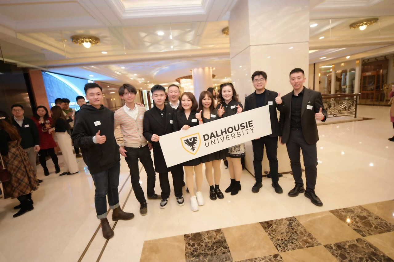 A group of alumni hold a Dalhousie sign at an event in a hotel lobby.