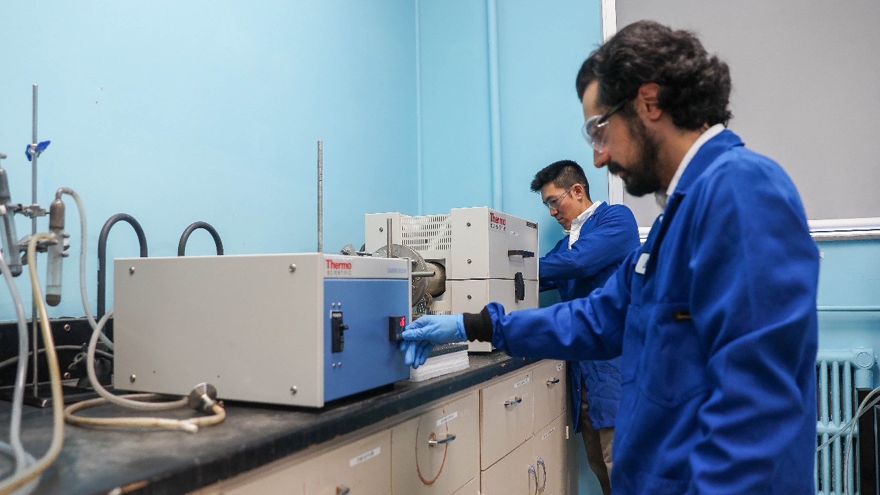 Two researchers wearing blue lab coats and safety glasses operate machinery in a lab.