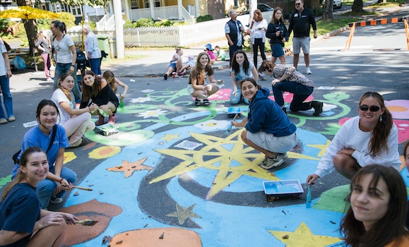 A group of young people sit on the street painting a brightly coloured mural.