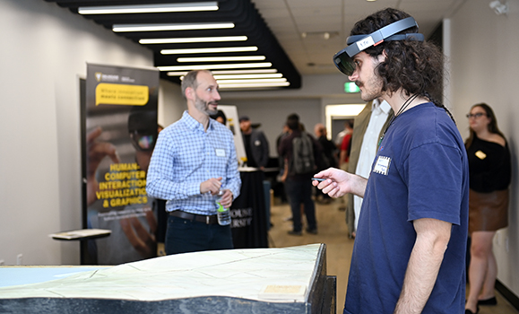 A man wears tech goggles while interacting with a demonstration at an event with other attendees in the background.
