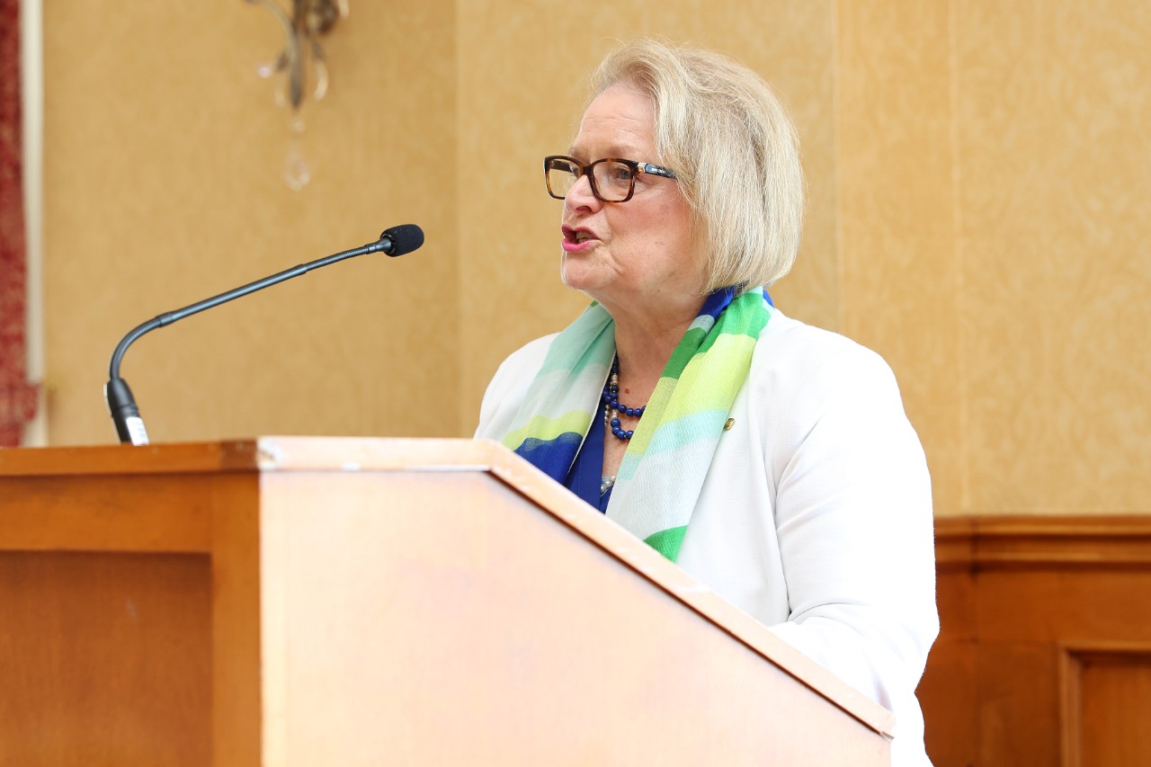 A woman with light blond hair wearing a white jacket and blue and green-patterned scarf stands behind a podium speaking into the microphone.