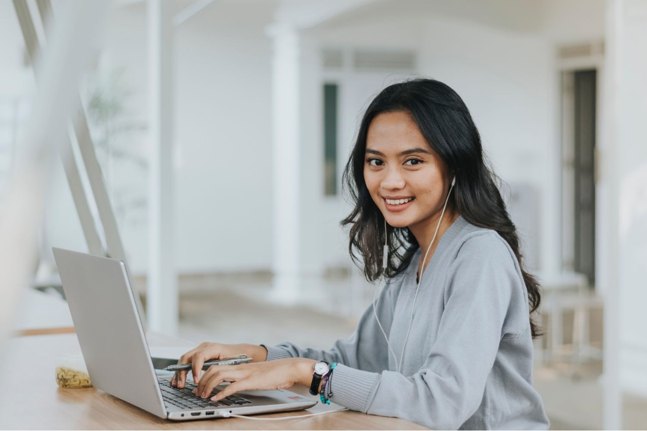 A female-presenting person in a light grey sweater is smiling while using a laptop and headphones in a bright, open indoor space.
