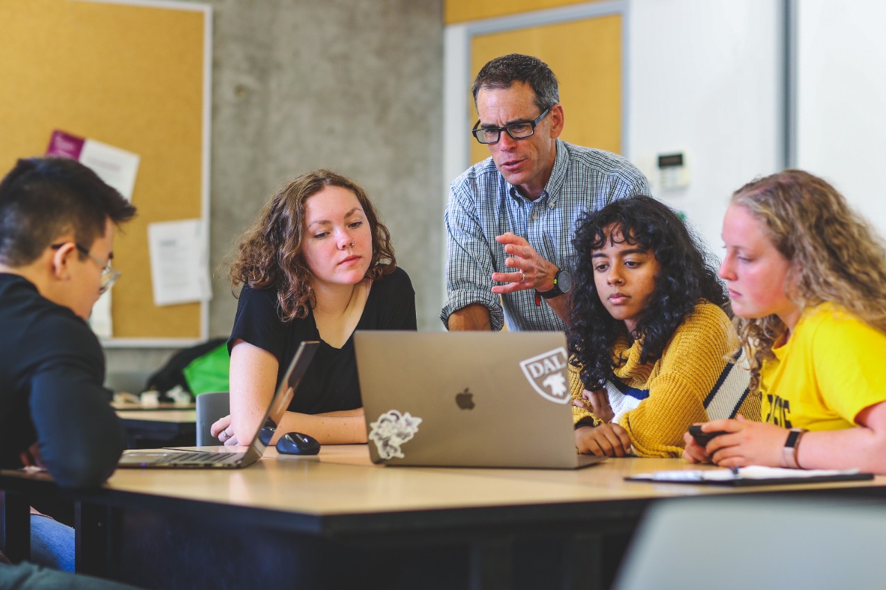 Four students sit around a desk and a fifth person standing inbetween them making an explanation.
