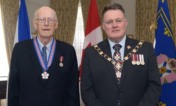 Dr. Allan Marble and Govenor General Michal Savage stand together smiling slightly adorned in their medals and suits with flags in the background. 