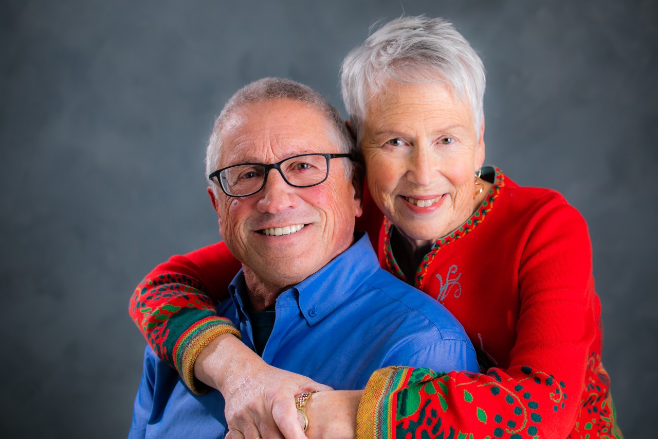 A woman with short grey hair hugs a man wearing glasses and a blue shirt