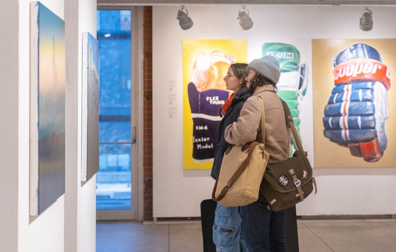 Two people viewing paintings on display gallery-style, dressed for winter and carrying satchels.
