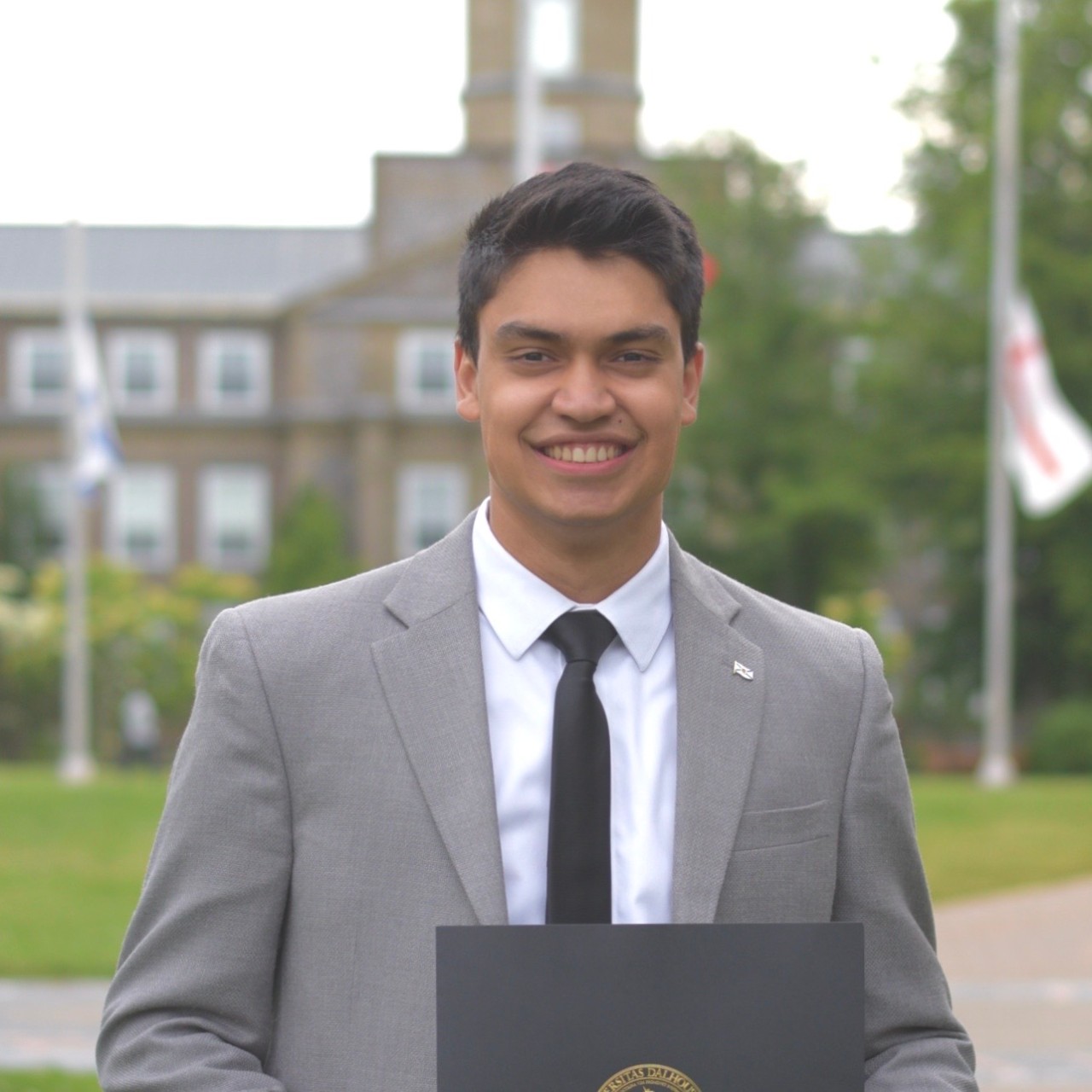Drabble dressed formally, smiling and holding a black folder on campus. 