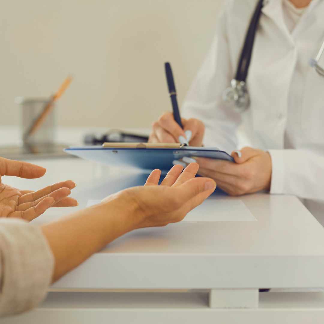 The hands are shown of one person open and gesturing while another in a lab coat and stethoscope on their shoulder writes on a clipboard. 