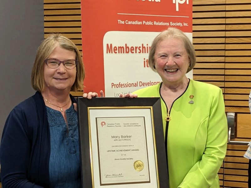 Barker is smiling and holding a framed certificate of her award alongside another women in front of a banner at an event.