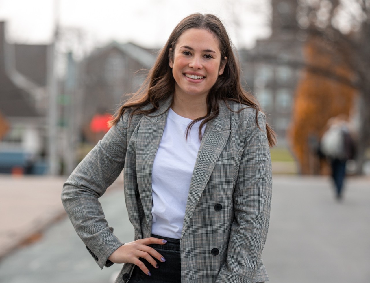 Kohn is standing in the street dressed business casual and smiling with her hand on her hip.