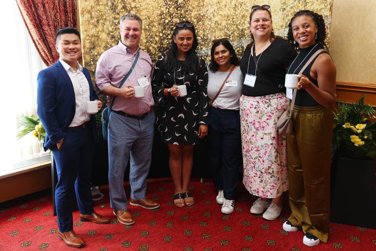 A group of 6 people are posing with white coffee mugs at an event.