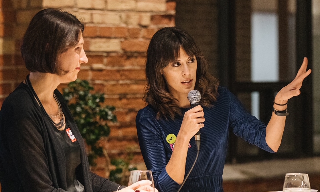 Two women are at a podium in front of an interior brick wall at an event where one is speaking into a microphone while the other looks on.