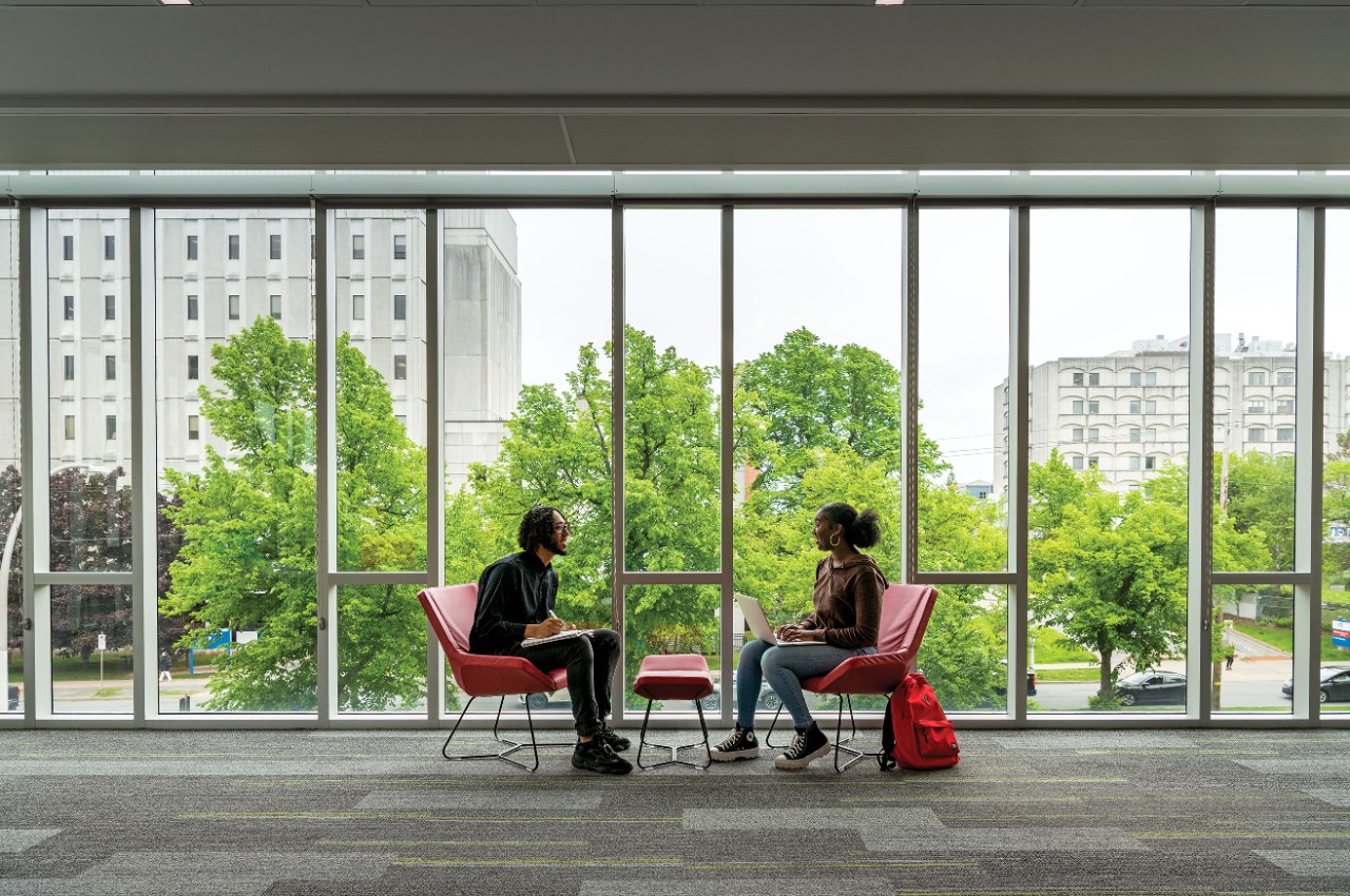 Students sit in red chairs facing each other. Large windows in the background frame a view of greenery and city buildings.