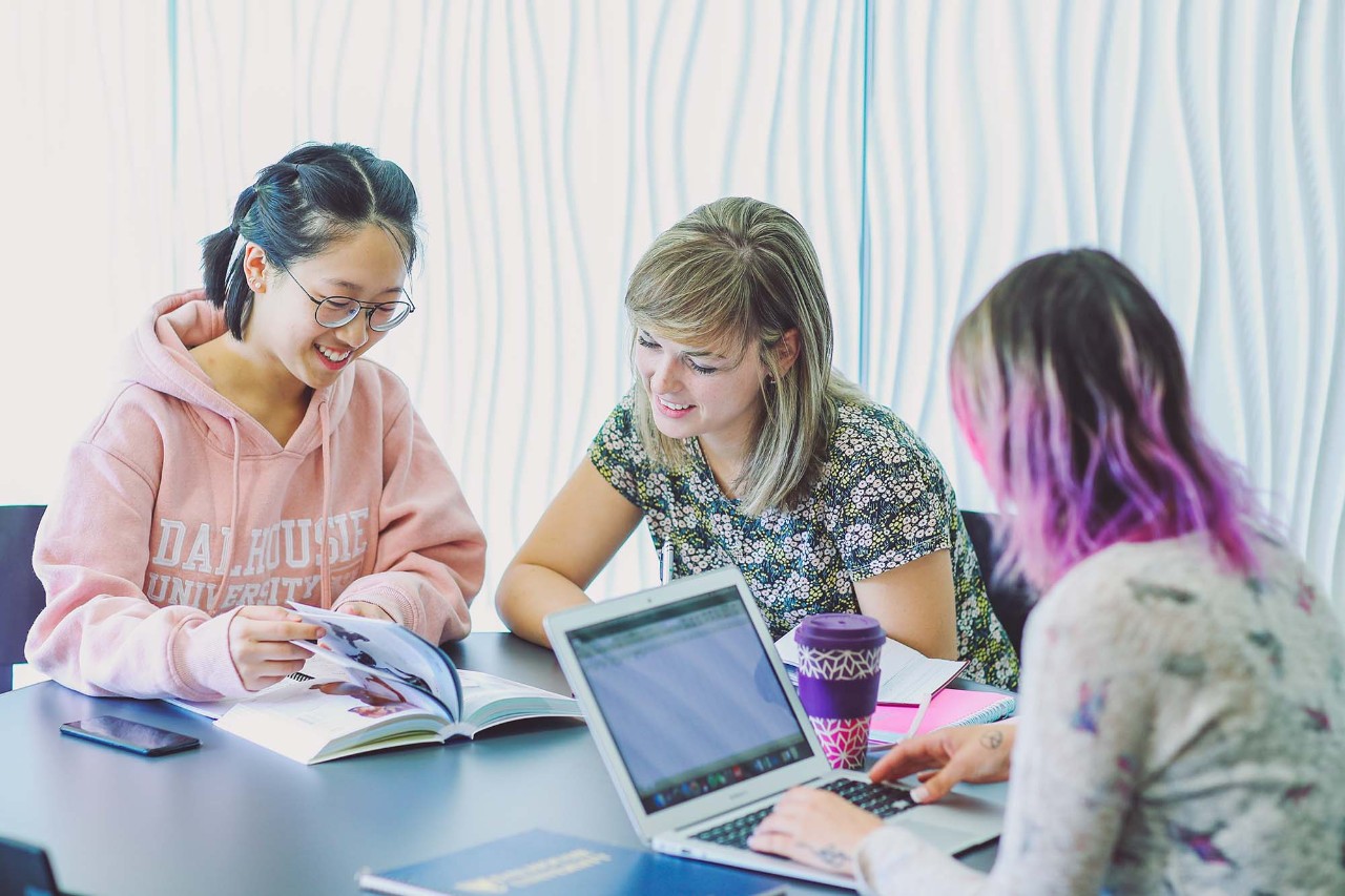 Three students around a table. Two students facing the camera are reviewing something in a book. 
