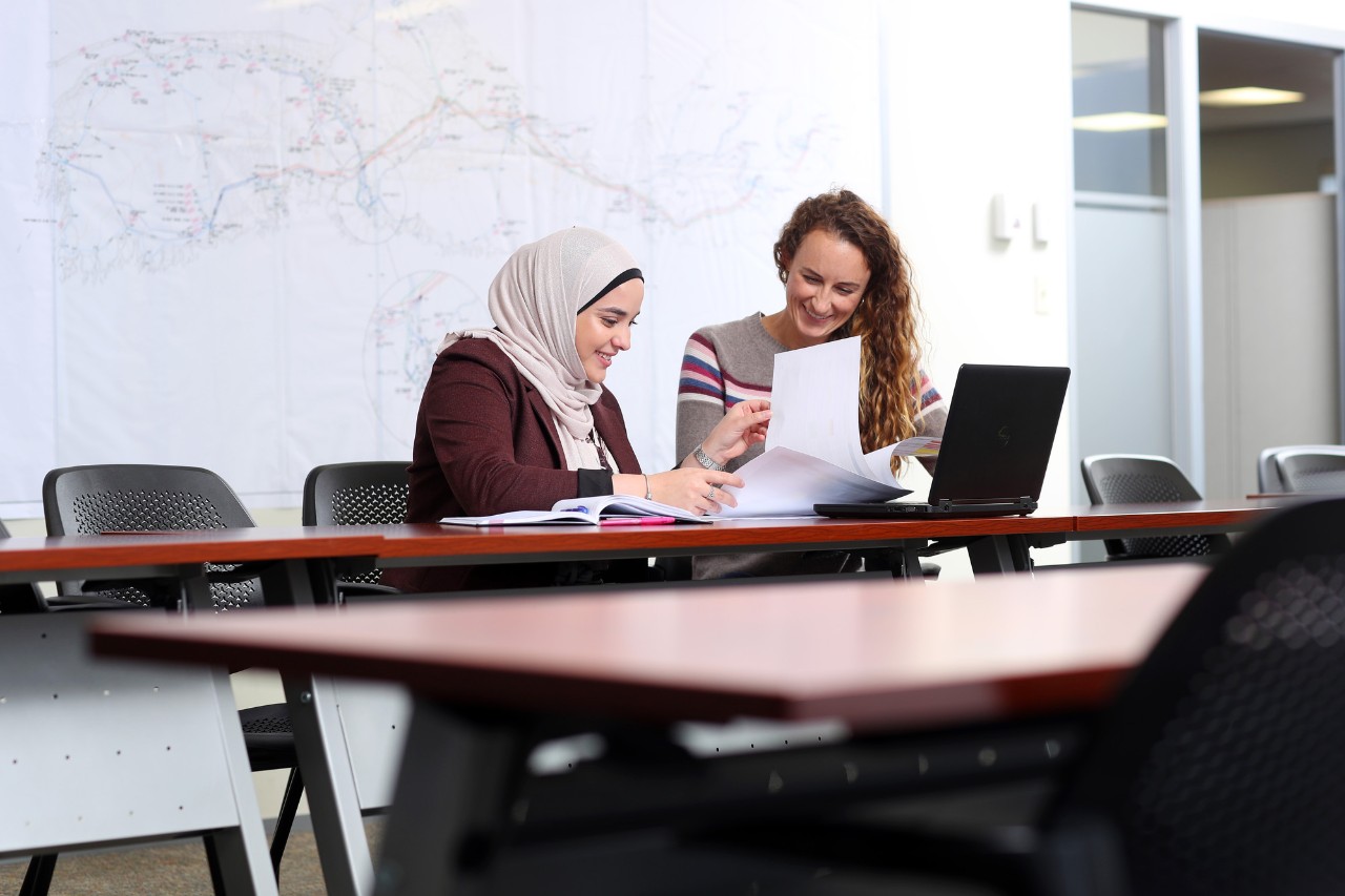 An instructor and student collaborate on a project while seated at a long desk.