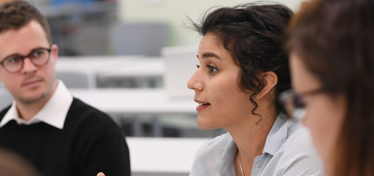 Two people are observing one person wearing a long sleeve pale blue shirt rolled up to the elbows speaking.