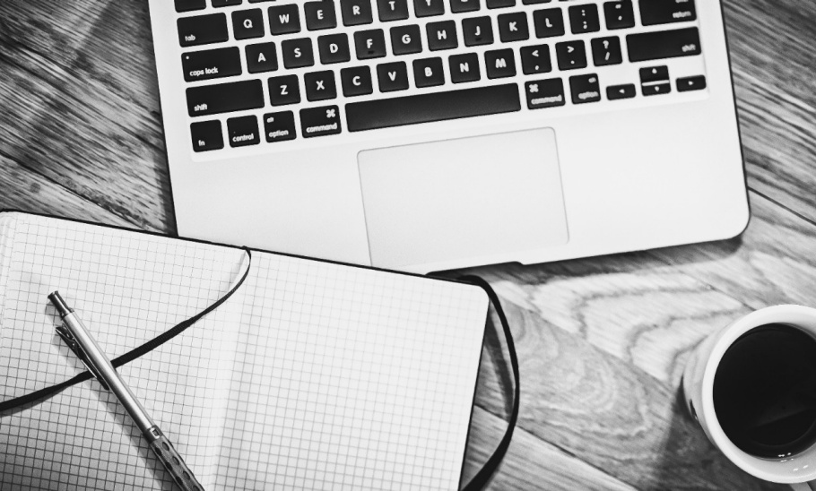 Black and white photo of a laptop, a notebook and pen, and a cup of coffee.