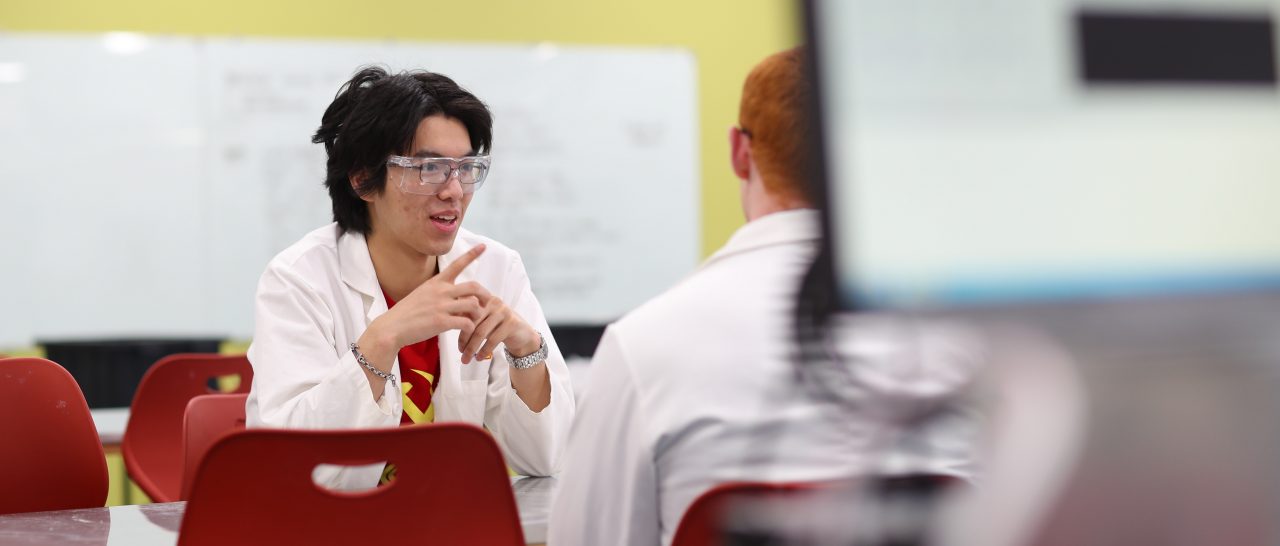 Two people are seen talking, both wearing lab coats, sitting at a table with red chairs.