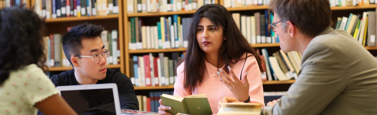 Four students sit around a desk with books and a laptop with stacks of books behind them.