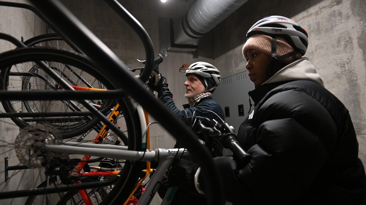 Two cyclists wearing helmets hang their bikes on an indoor mounted bike rack.