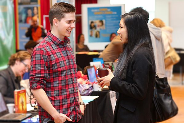 Two people are in conversation at a career fair with tables set up in the background and banners.  
