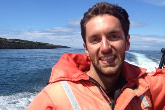 A close-up of alum Will Burt on a boat with the wake and land in the background, smiling and wearing a heavy orange jacket. 