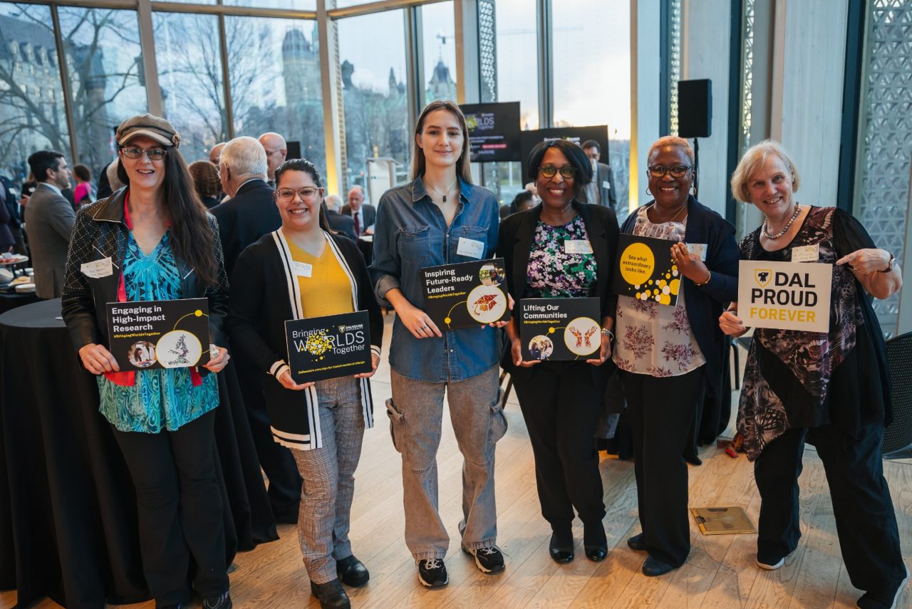 A group of six women hold signs with Dalhousie branding during an event in Ottawa with high floor to ceiling windows.