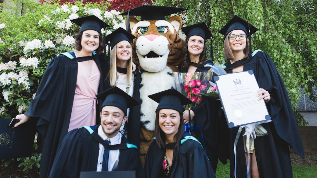 Six students in grad caps and gowns are posting with the Dal Tiger, also in a grad cap, in front of campus foliage. Three students are holding their degrees and one student is holding a bouquet of roses.