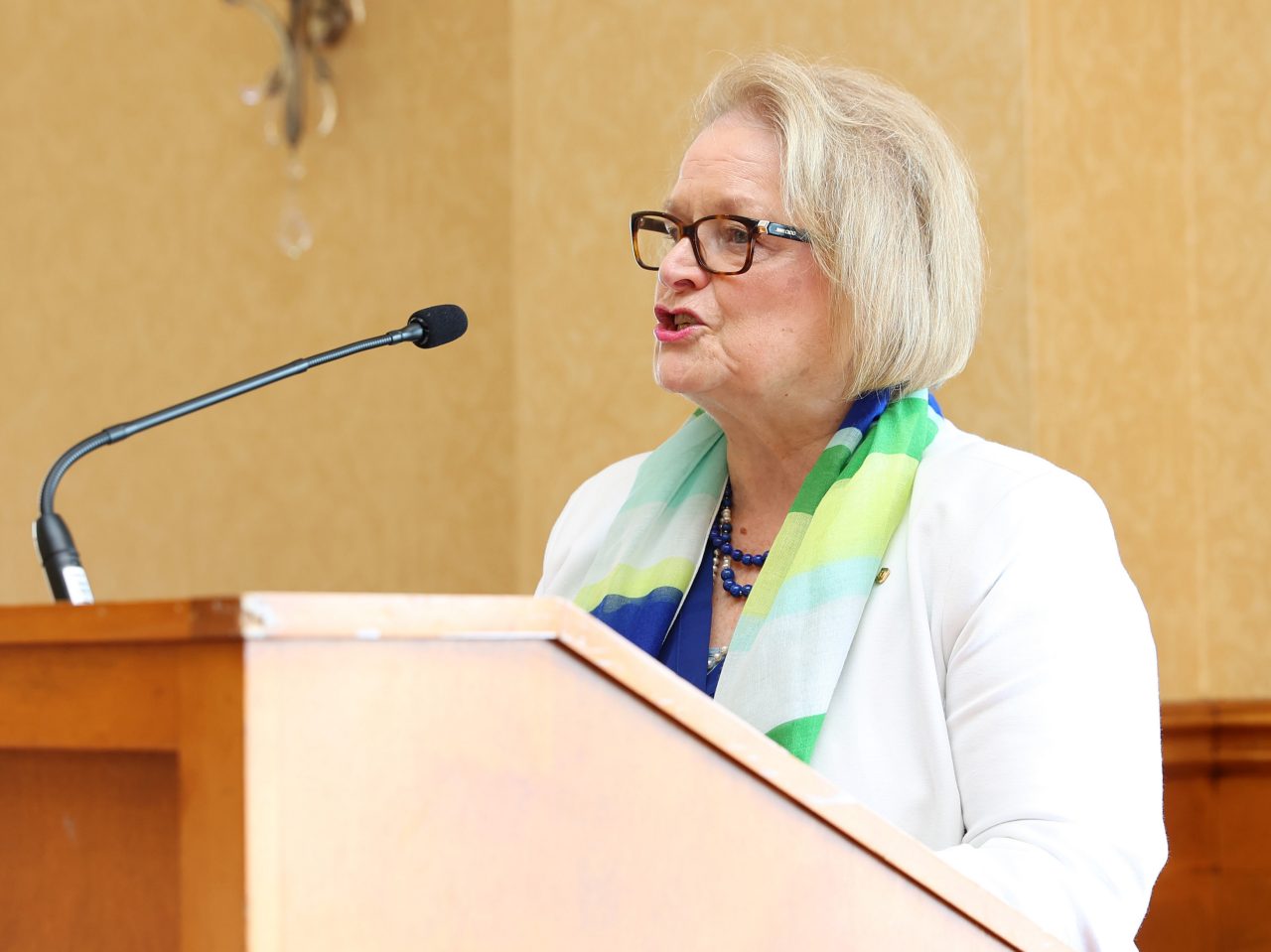A woman with light blond hair wearing a white jacket and blue and green-patterned scarf stands behind a podium speaking into the microphone.