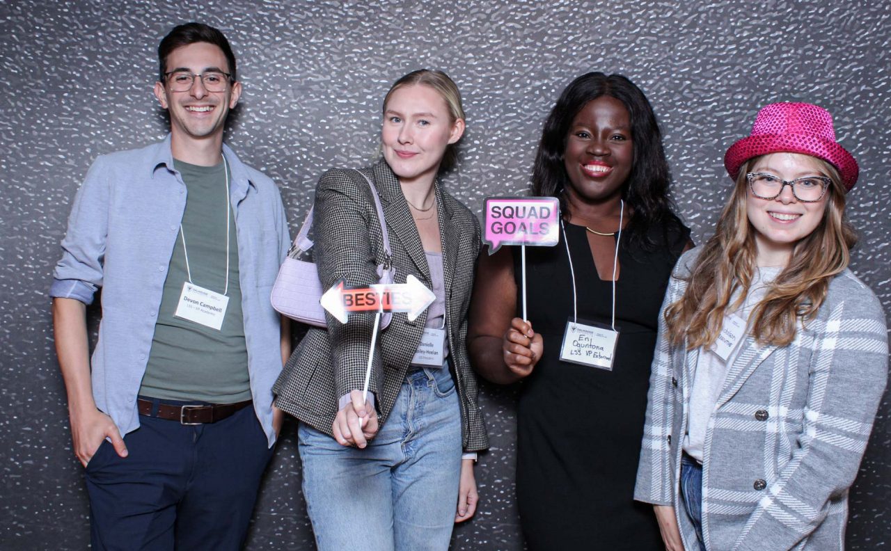 Four people stand smiling against a textured grey backdrop. They wear nametags and hold 2 photobooth signs on sticks.