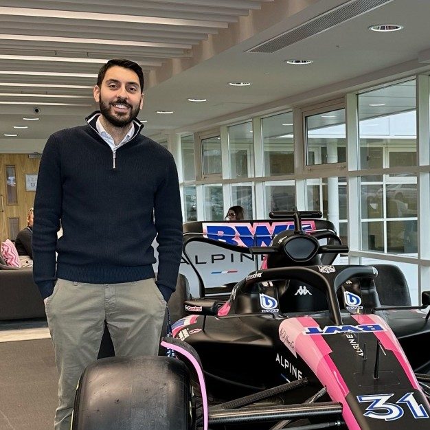 Ahluwalia stands smiling next to a BWT racing vehicle indoors next to a wall of windows.