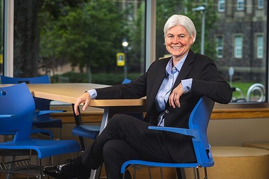 A woman wearing a black suit and blue dress shirt sits casually in a chair