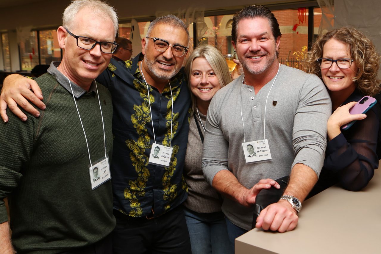 A group of 5 people stand close together at a casual get together smiling and wearing nametags on lanyards.