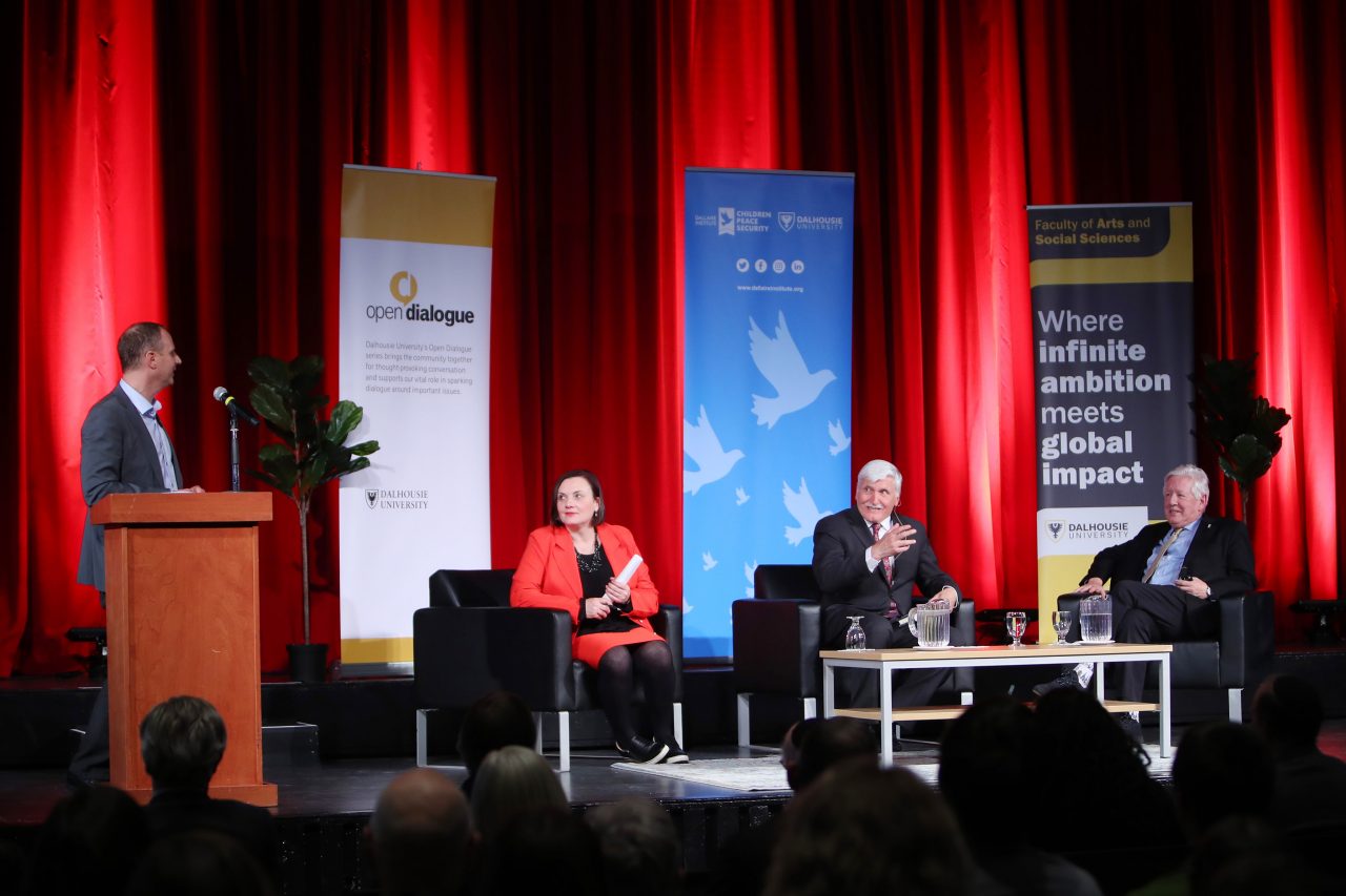 Three people sit on stage with banners red curtains in the background while another person stands at the podium addressing them.