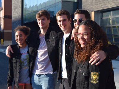 A group of engineering students stand with their arms around each other and smile in front of the IDEA building. 