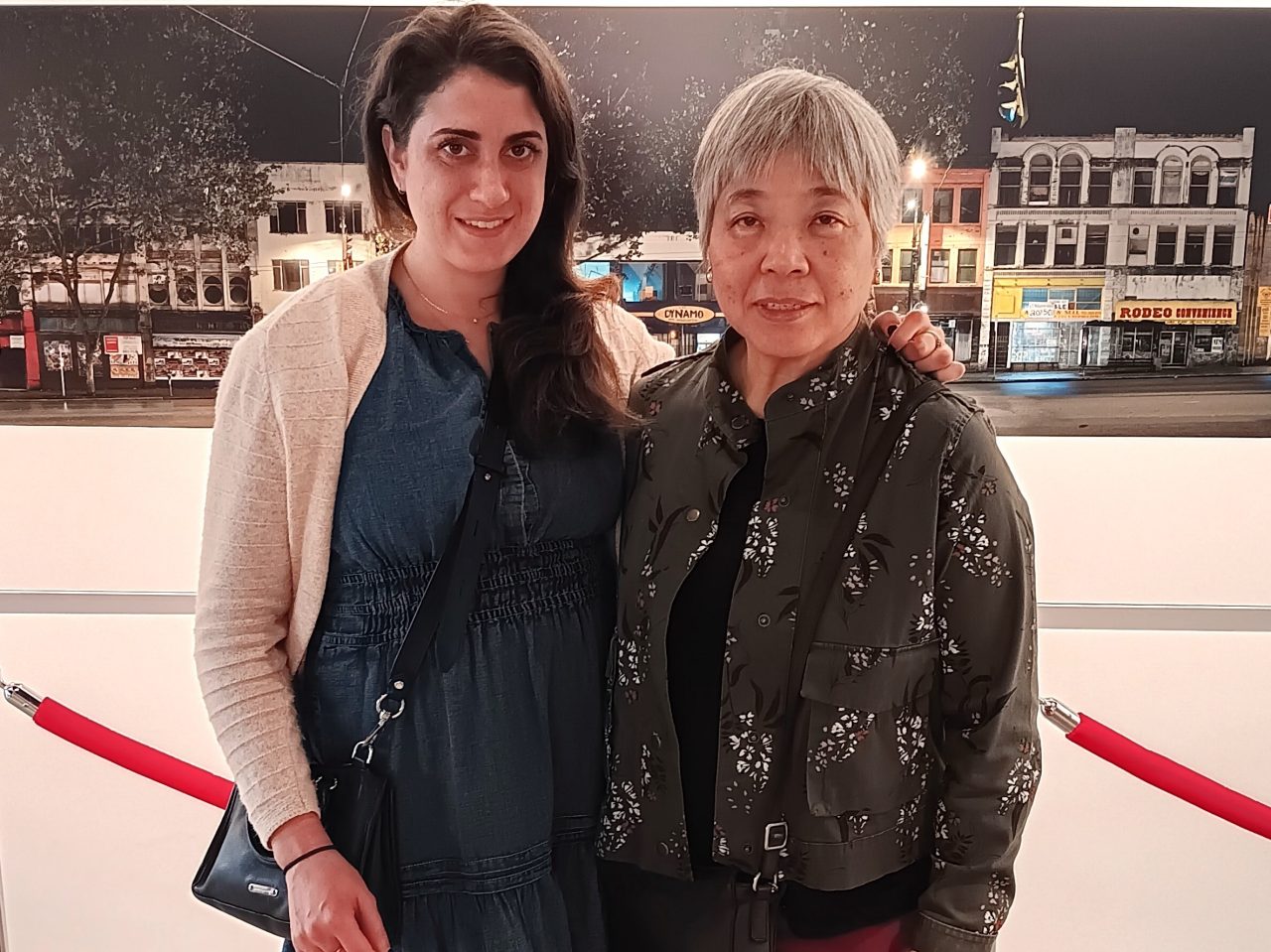 Two woman dressed casually stand together indoors in front of a photograph of a streetscape.