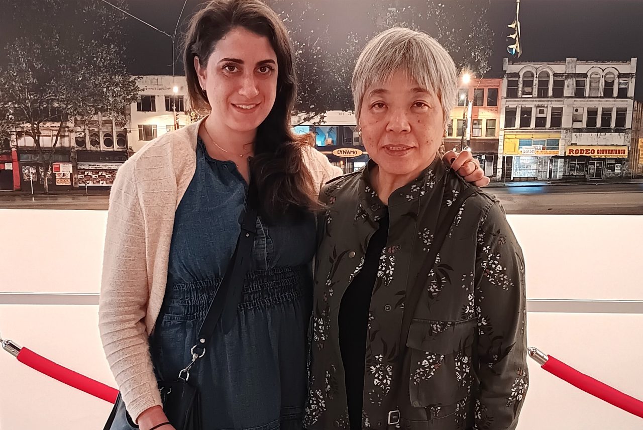 Two woman dressed casually stand together indoors in front of a photograph of a streetscape.