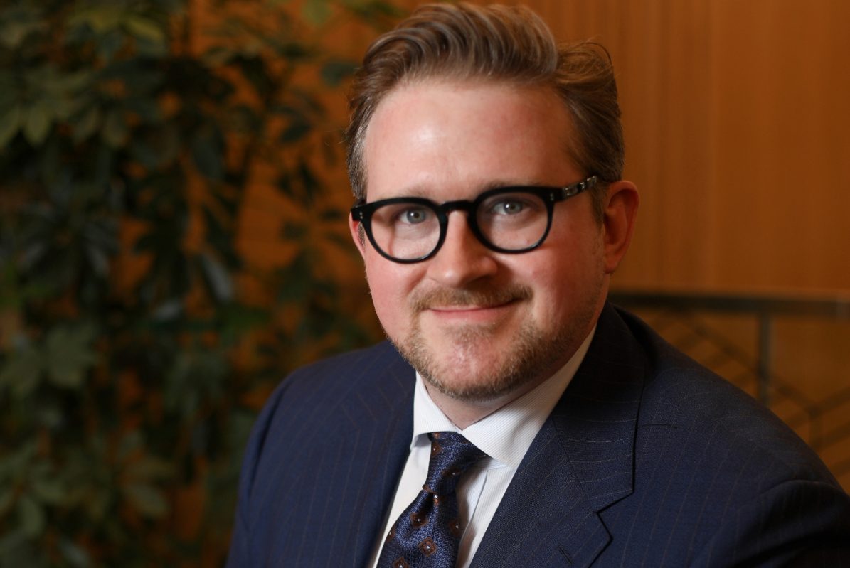 A headshot of a seated man wearing glasses and a blue business suit